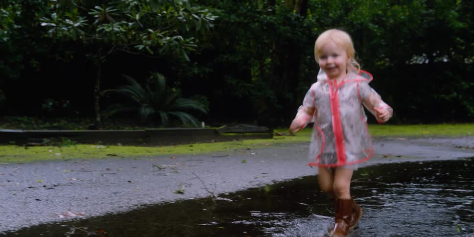 Toddler blonde girl with a transparent raincoat and brown boots runs through a puddle in the park