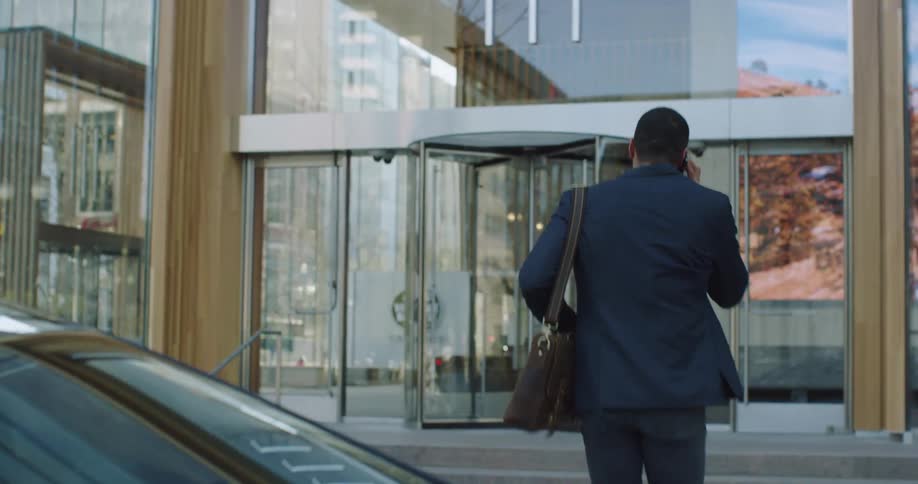 Business man talking on the phone exits a car to enter a building in the city