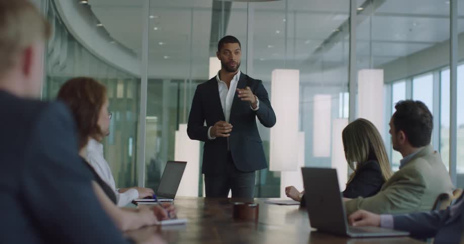 A man stands at the end of a table in a conference room giving a presentation in front of his coworkers during the day