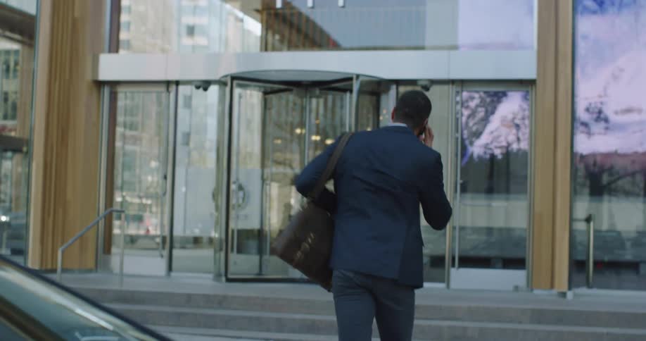 A man gets out of a car while talking on the phone and walks inside a large city office building