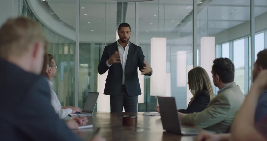 A man standing in front of a table speaks in front of coworkers who are sitting and listening to him speak inside of a conference room