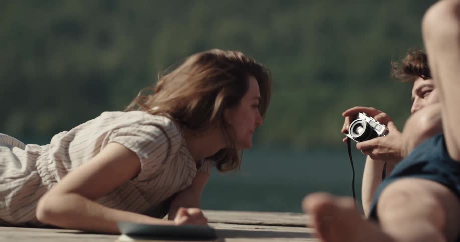 A man lying on a dock photographs his girlfriend as she poses and makes faces