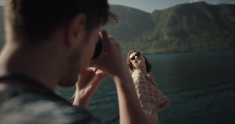 A man uses a small camera to take photographs of a young woman posing on a pier on a river with a shawl