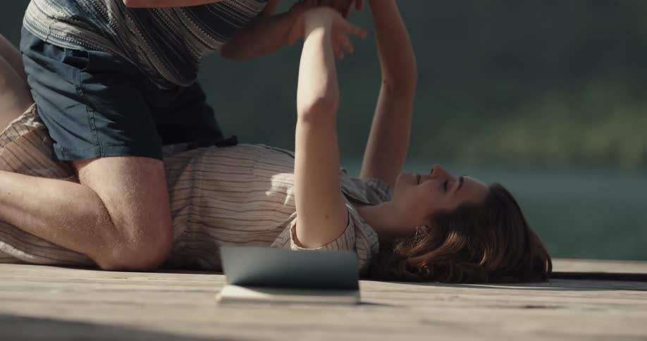 A woman lying on her back on a pier next to a river makes funny faces at a man kneeling over her with a camera