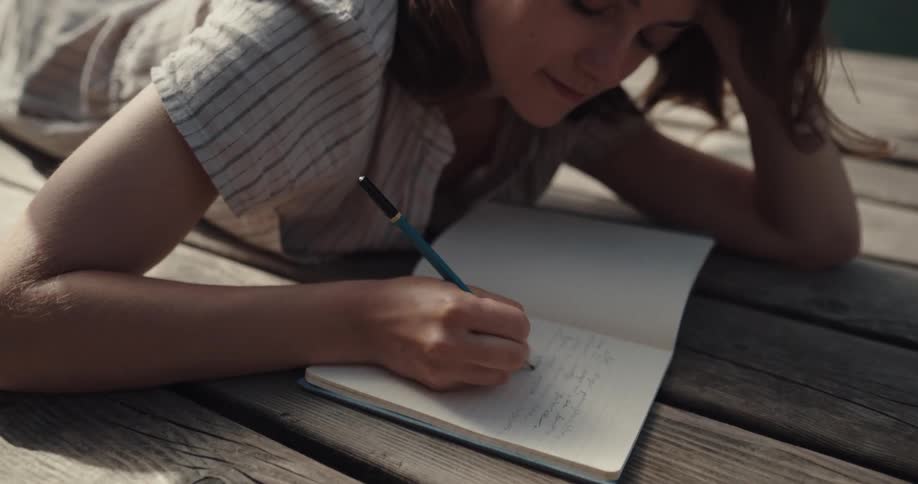 A woman smiling while writing in a diary lakeside on a wood dock