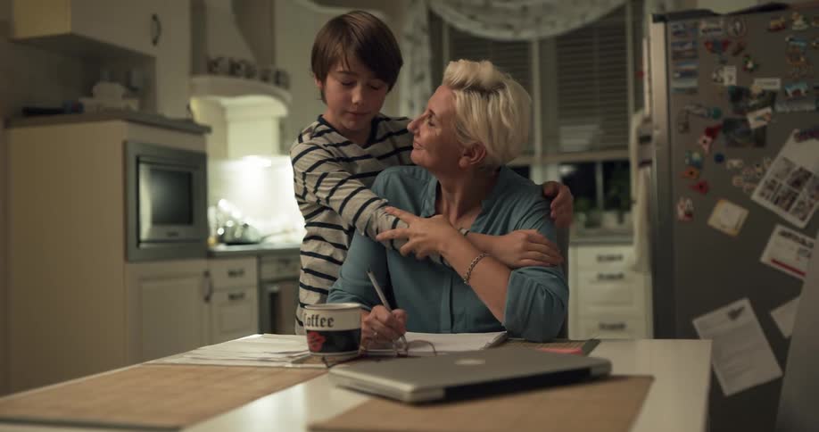 A young boy hugging his mother while she works at a kitchen table