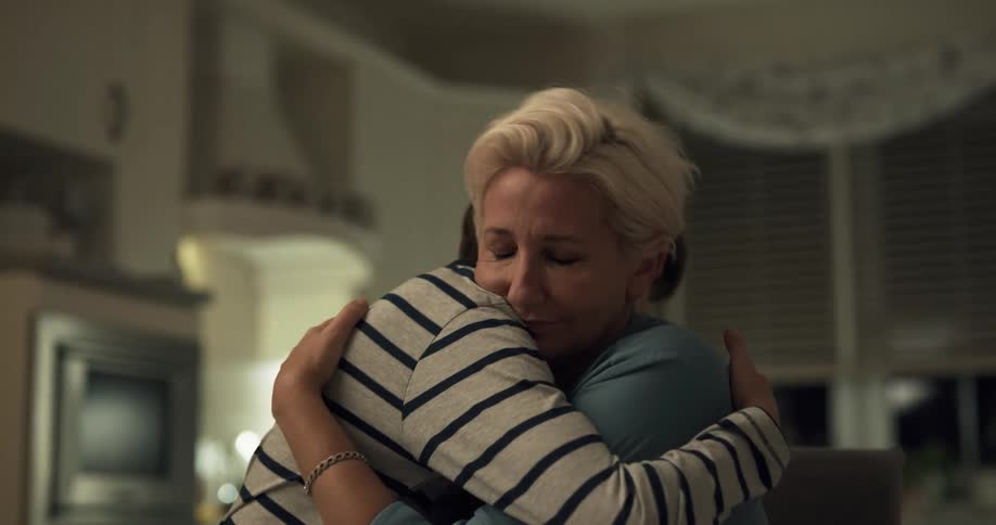 An elderly woman sits looking at a paper and her grandson comes up to give her a hug