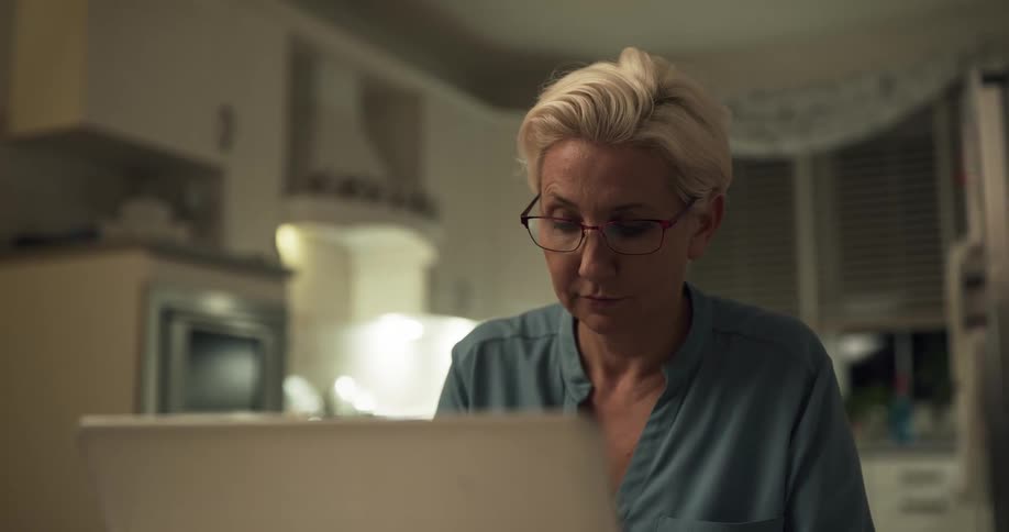 A woman sits at a table indoors next to a kitchen and works on a silver laptop computer at night