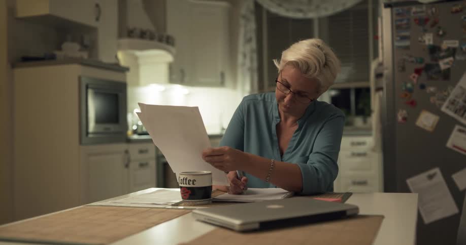 A woman sits at a kitchen table looking at paperwork