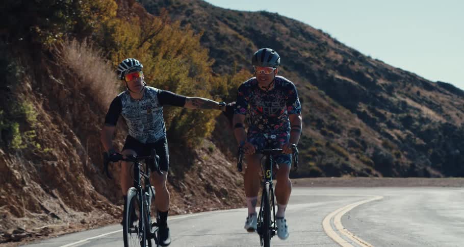 Two cyclists pedal bicycles along road that hugs slopes under clear blue sky