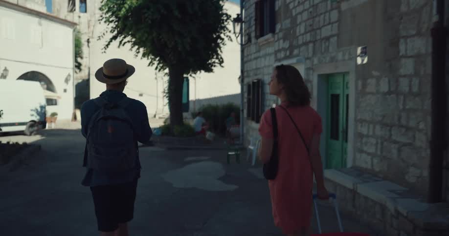 A man walks next to a woman pulling a suitcase as they both look around down an alleyway next to a building during the day