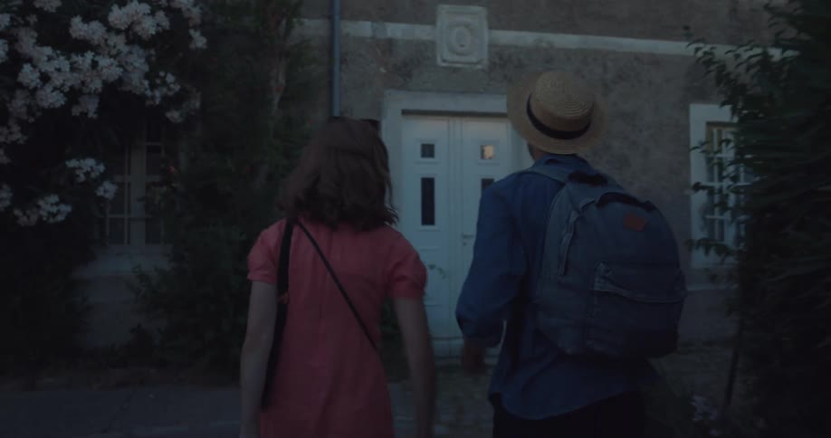 A man and woman walk to a building front door with their luggage