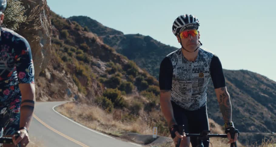 Two heavily tattooed cyclists ride along the edge of a curvy mountain road