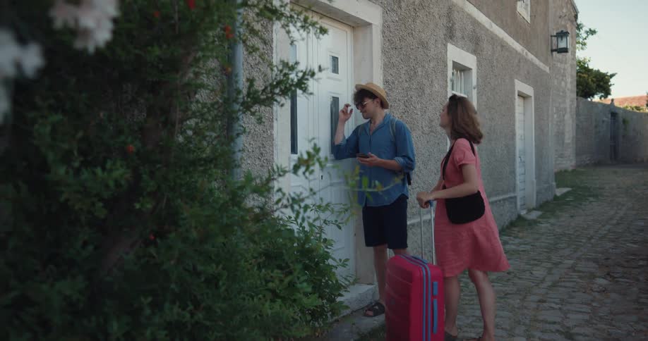 A man and a woman with travel gear walk up to a front door and knock during the day
