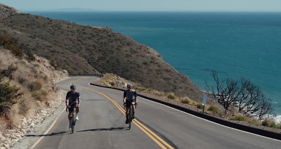 Two people are riding bicycles on a winding road next to an ocean