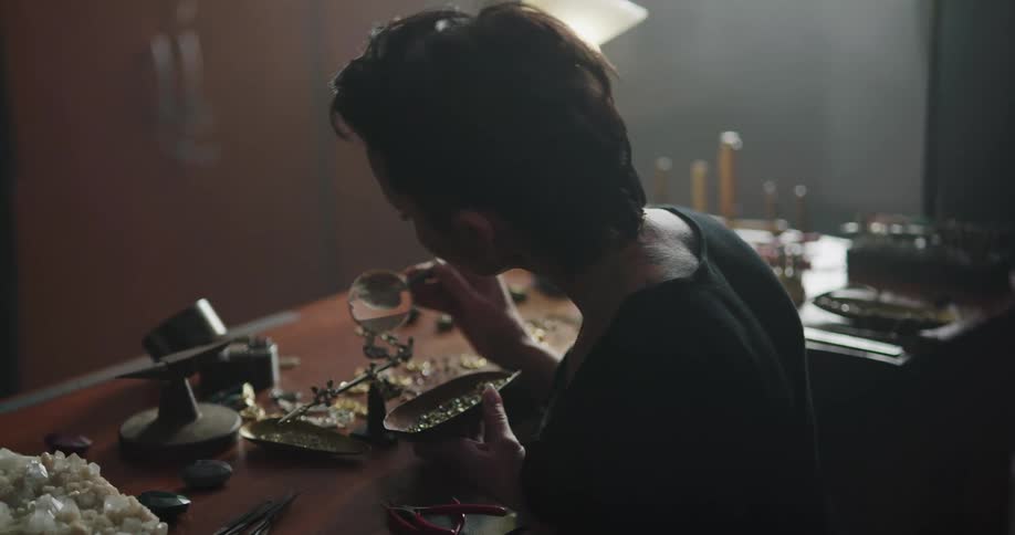 A female jewelry maker sits at a desk and fits gemstones into a setting that she is working on