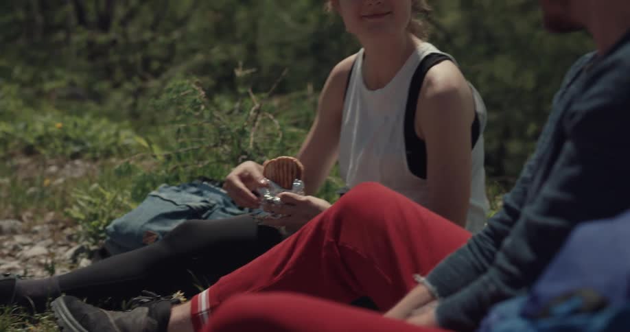 A man and woman sit in a forest and the woman offers a bite of her sandwich to the man