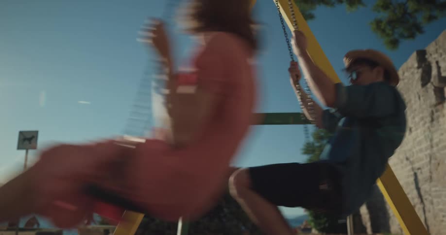 Man and woman swing on a swing set near a brick wall under a blue sky