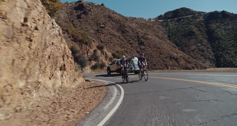 Two people are riding bicycles on a mountain road as a truck goes past them