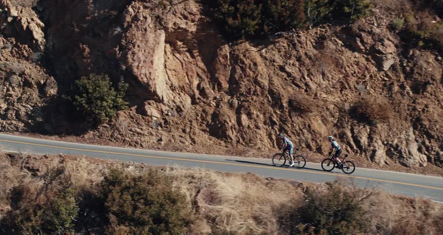 Two people riding bicycles on the highway near a cliff with plants and trees