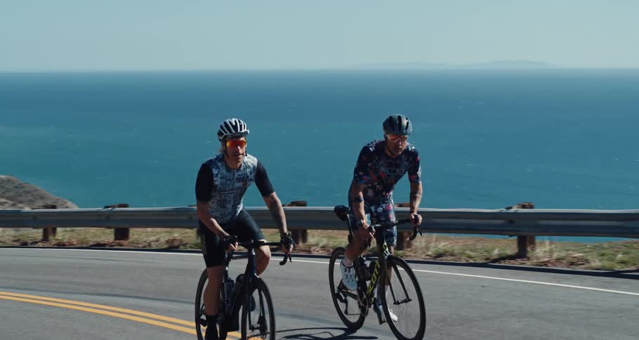 Two men cycling up a road next to a hill overlooking the ocean during the day