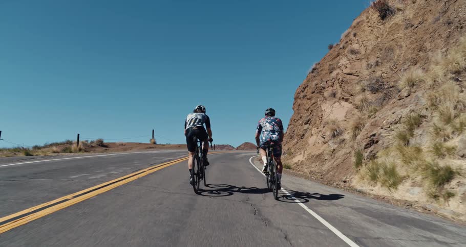 Two cyclists pedal bicycles along highway among hills as bird flies overhead