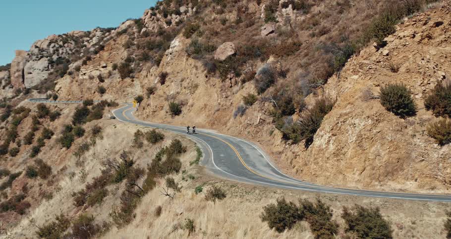Two people are riding bicycles on a winding mountain road with mountains in the distance