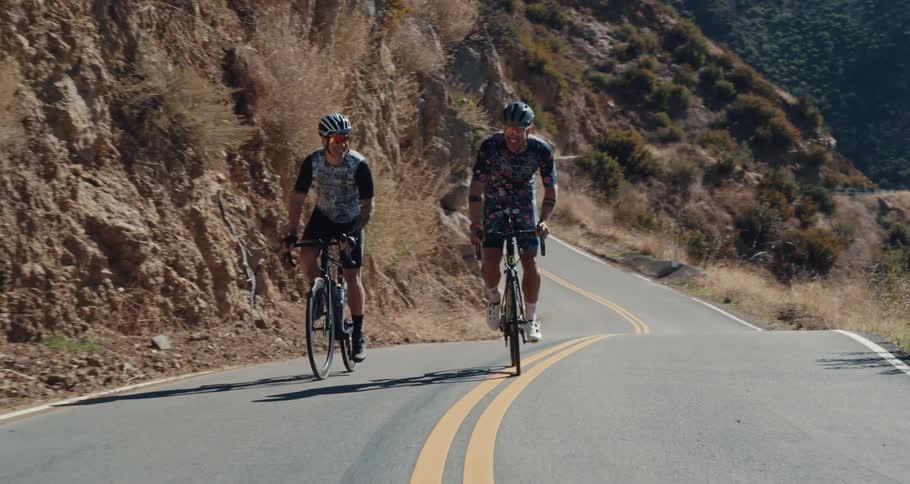 Two cyclists wearing helmets pedal down winding road hugging mountain slope