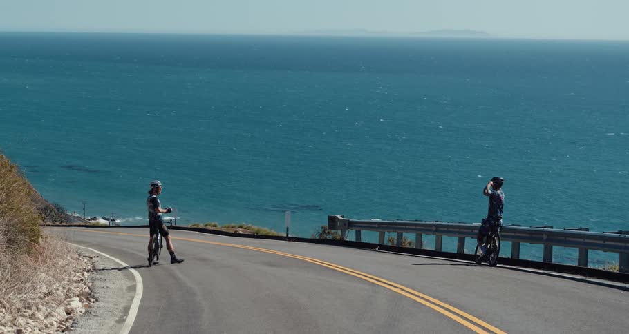 A cyclist stands in the middle of the road to take a picture of another cyclist with an ocean background