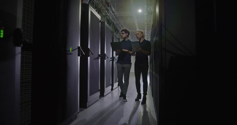 Two workers holding laptop computers observe and point at computer servers in a server room