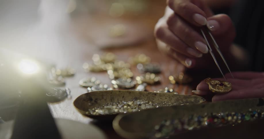 A female jewelry maker carefully picks up small gem stones and places them into a golden pendant