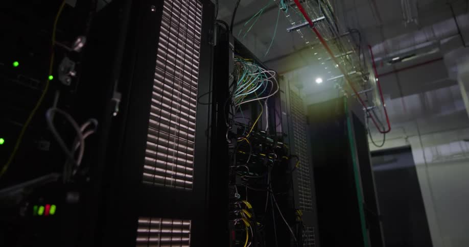 A room with server racks with wires and lights with a wall and door in the background
