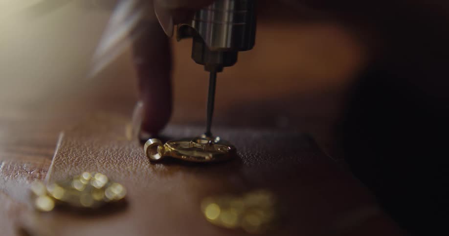 A woman uses a tool to make a puncture in a small gold jewel on a table