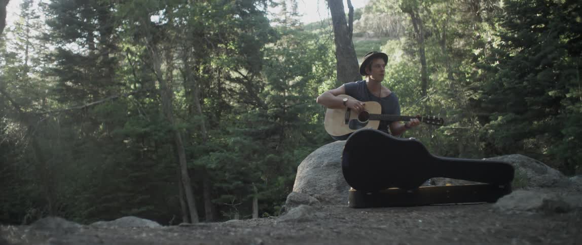 A man is sitting on a rock in the woods playing a guitar