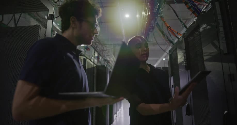 A woman holding a tablet talks with a coworker in a data center in an aisle of servers and wires