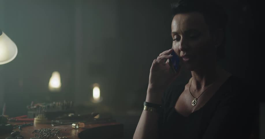 A woman with a necklace in a room talking on a smart phone sitting at a desk