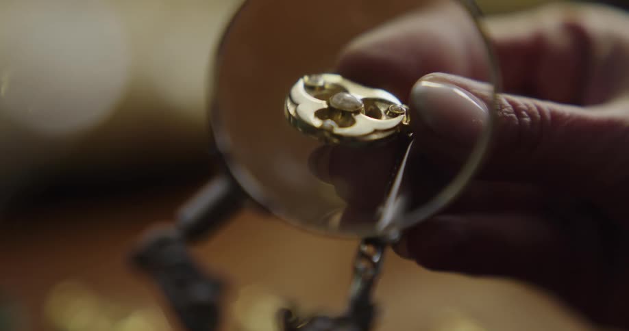 A woman moves around a small piece of gold jewelry under a magnifying glass