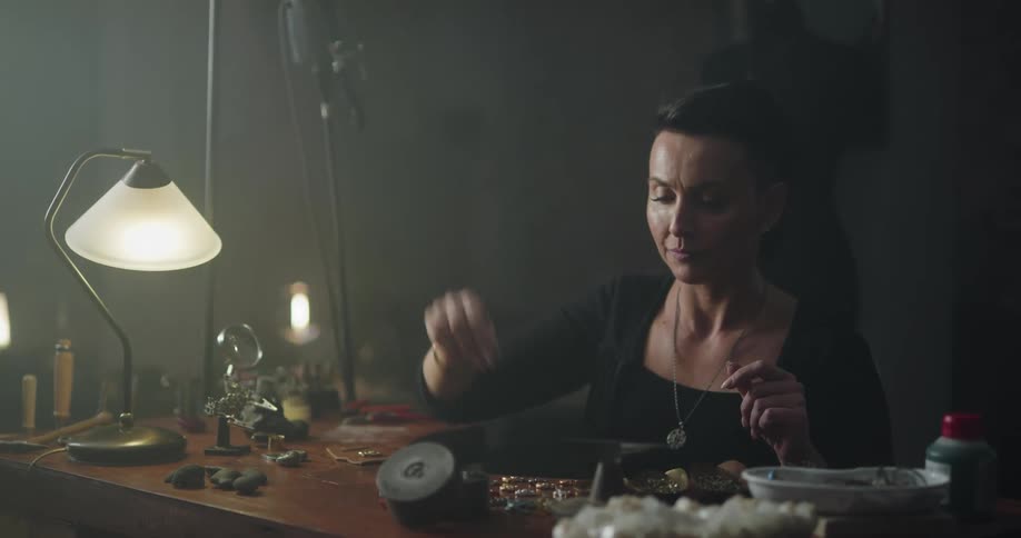 Jewelry artist works with beads sitting and table in dim room lit by desk lamp