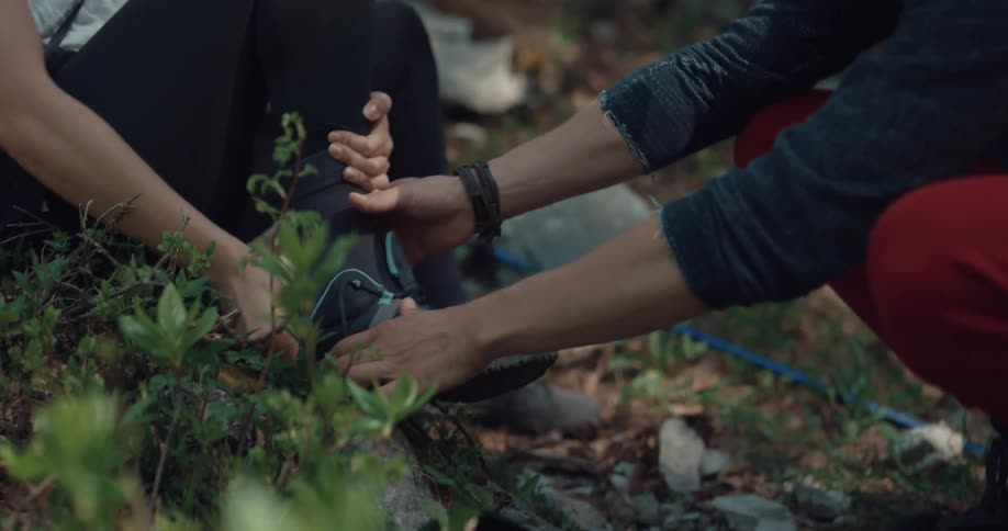 A man and woman in a forest surrounded by plants with the man removinga sneaker from the womans foot