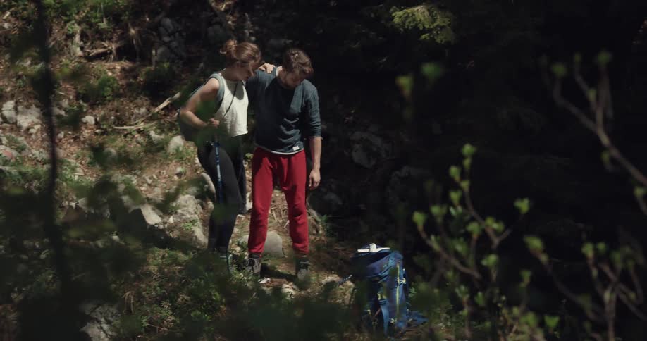 A male hiker helps an injured female hiker to her feet before retrieving his backpack from the ground