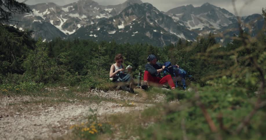 A man and woman empty their backpacks while sitting on the ground near snow capped mountains