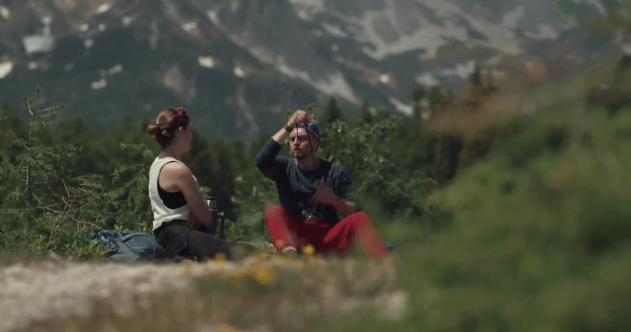 A man and a woman play fight over a small digital camera while sitting in grass in a forest during the day