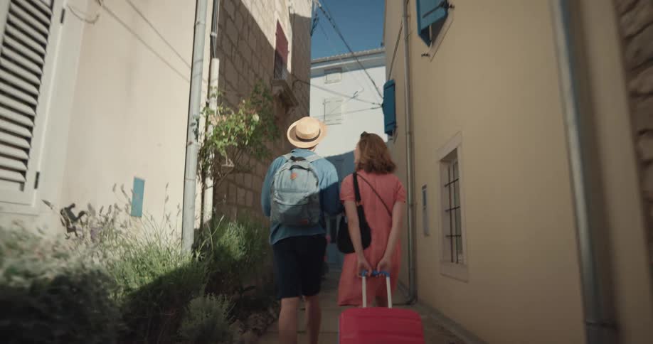 A man and woman look at a cell phone for directions as the woman pulls a suitcase down a narrow path