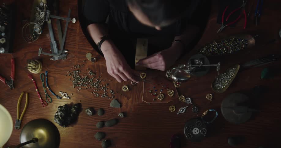 A female jewelry make sits at a desk with a wide assortment of gems and medal pieces in front of her