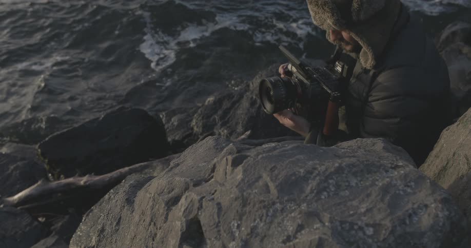 A man is photographing as he sits in rocks by the water