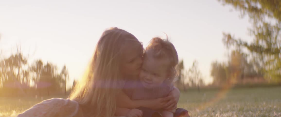 Two baby sisters hug on a grassy field with trees the sky and clouds in the background