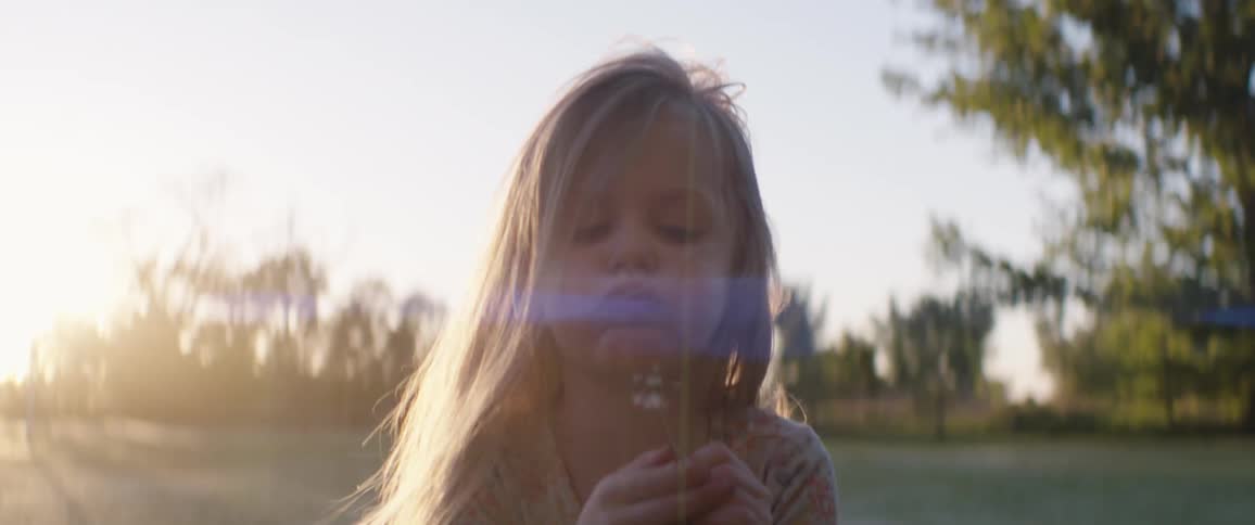 Blonde girl with long hair blows spreading dandelion into the wind while standing in field