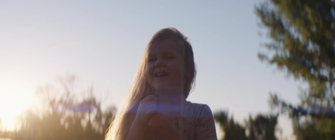 A little girl with long hair blows on a dandelion causing seeds to fly