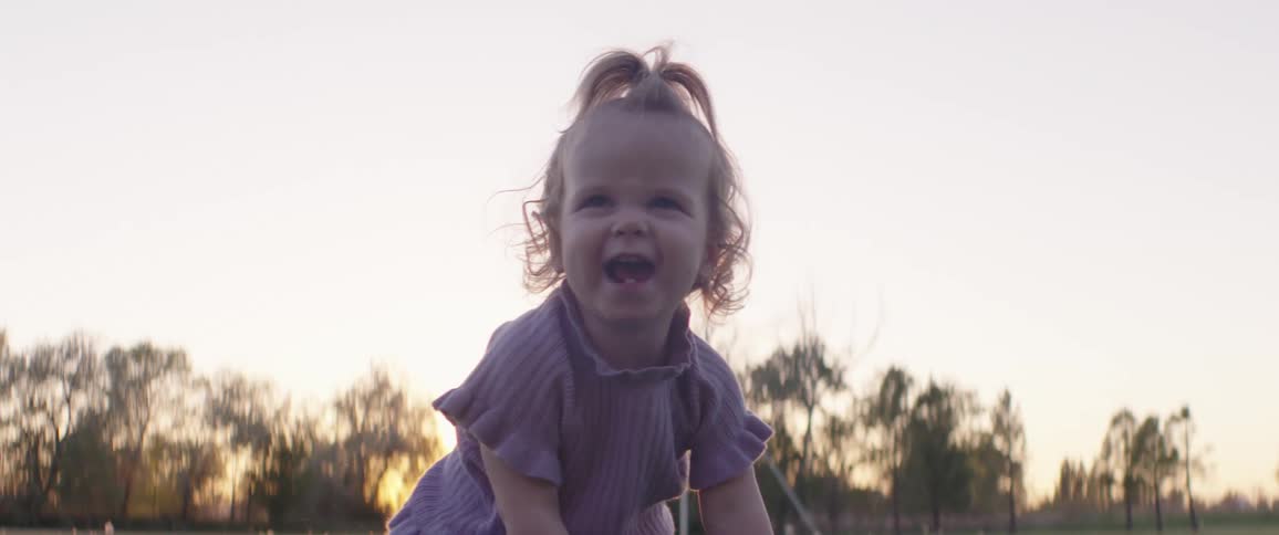 A little girl laughing on a grassy field with trees the sky and clouds in the background