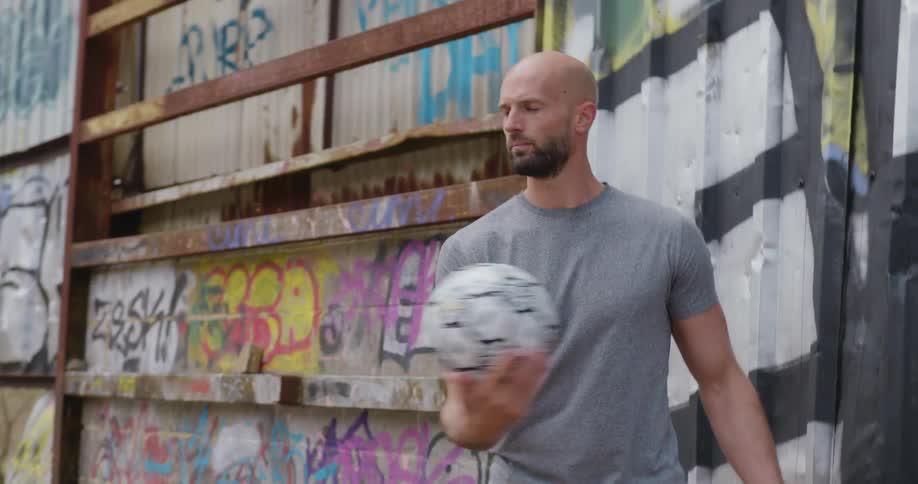 A man stands spinning a soccer ball on his finger with a graffiti wall behind him